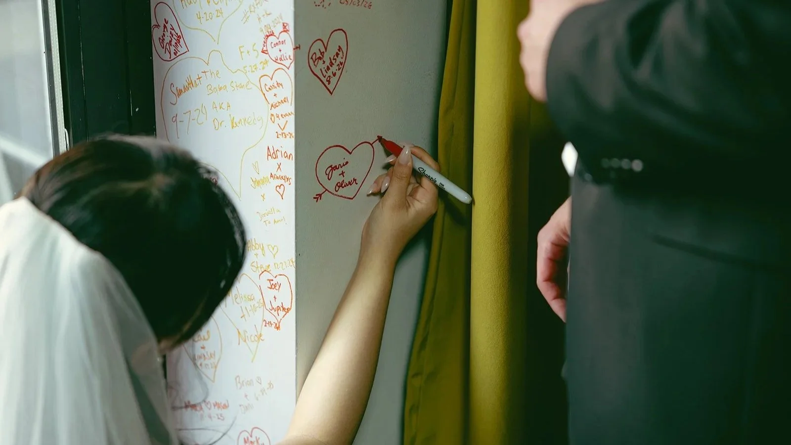  Two people sign a wall with a permanent marker. They are adding their names within a heart. 