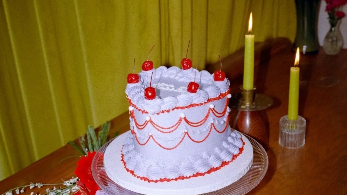  A light Purple Heart-shaped Lambeth-style cake with cherries on top sits on a bar top near green taper candles. 