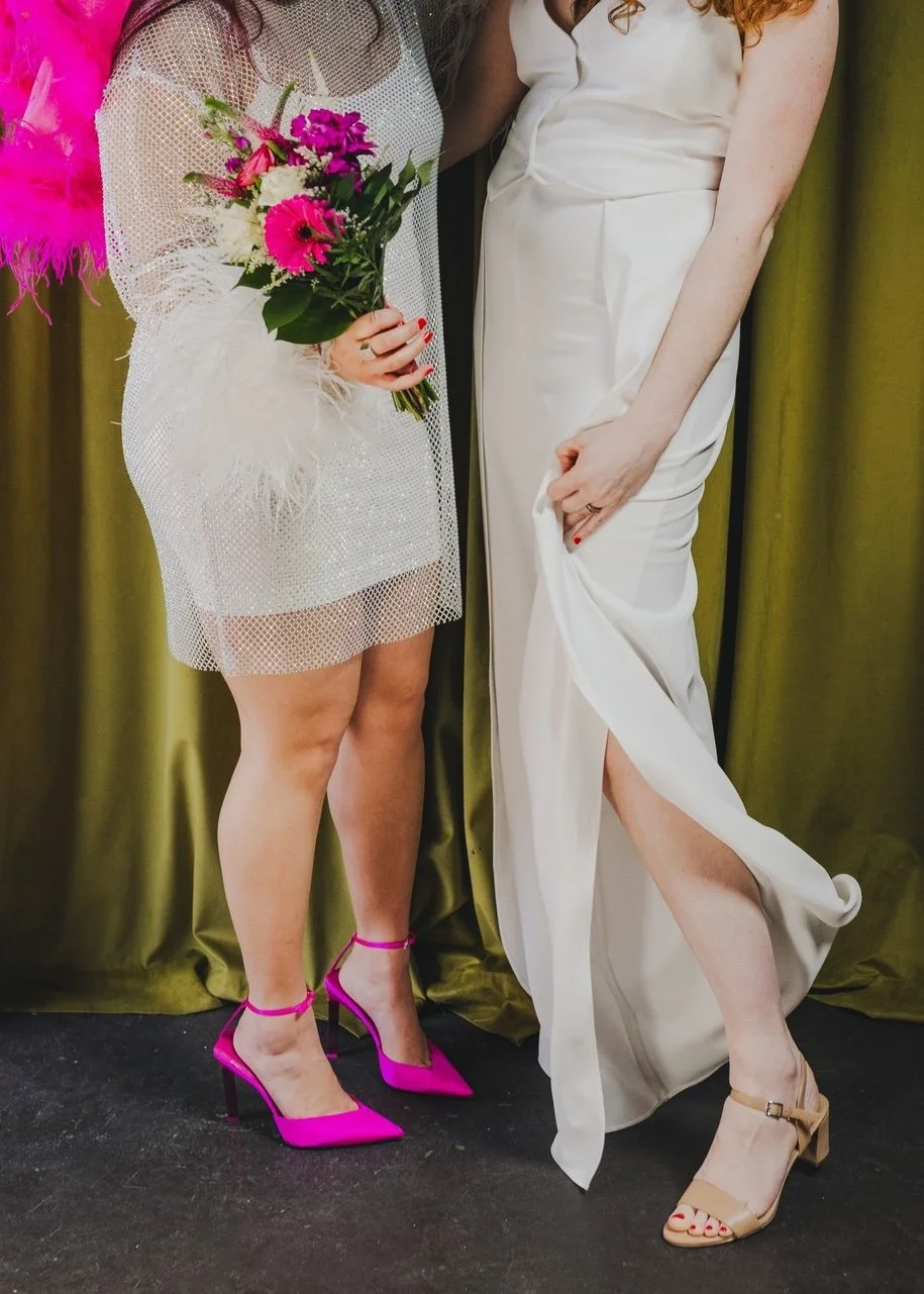 A couple stands side by side against a green curtain. One is wearing a short, sheer, sparkly dress with a hot pink veil and matching shoes. The other is wearing a white jumpsuit with a slit down the leg.