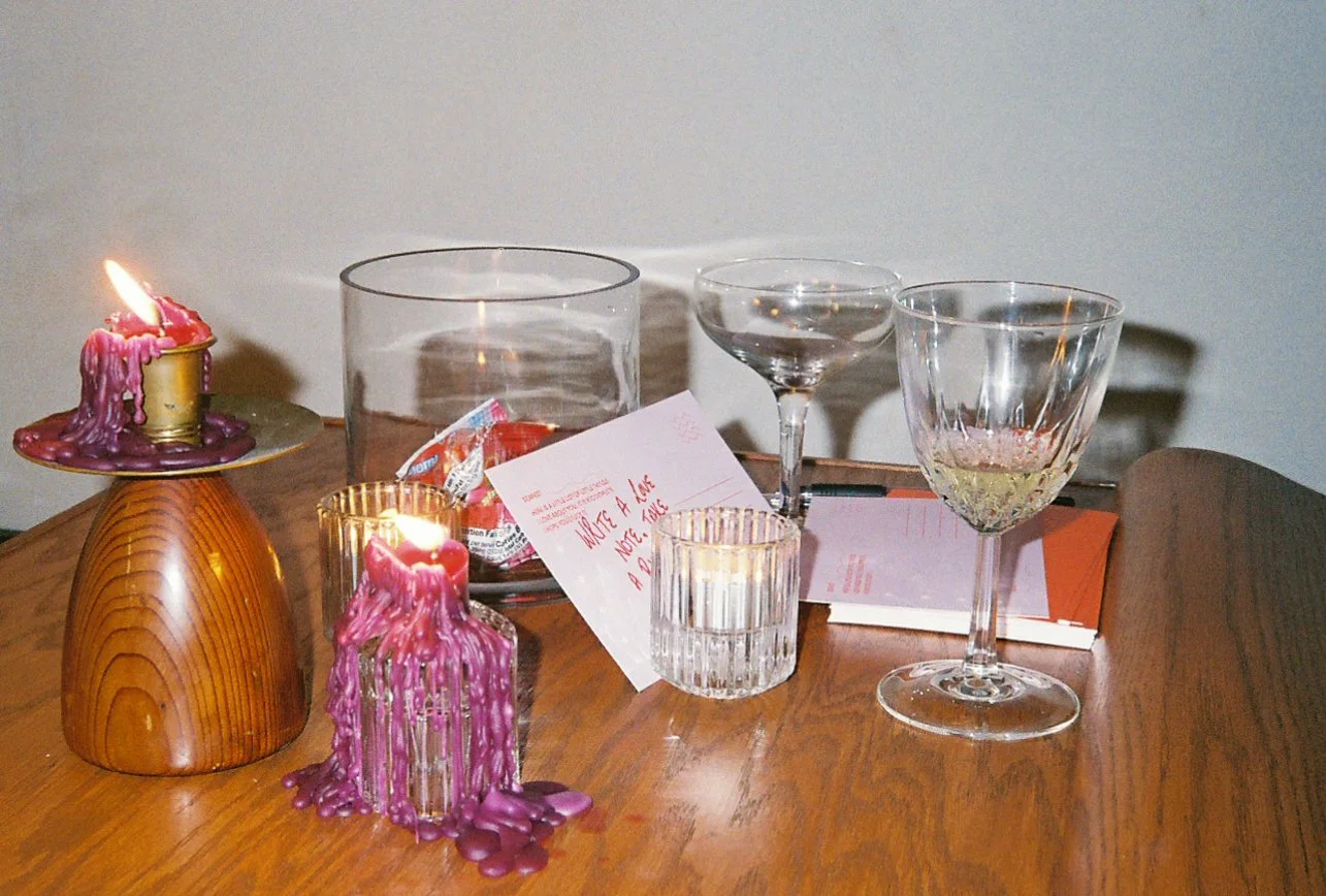 A wooden table with two taper candles melting beside a wooden candle holder, a clear glass bowl with red and orange wrapped ring pop candies, cocktail glasses, a small glass with a gold candle holder, and pink handwritten notes on postcards.