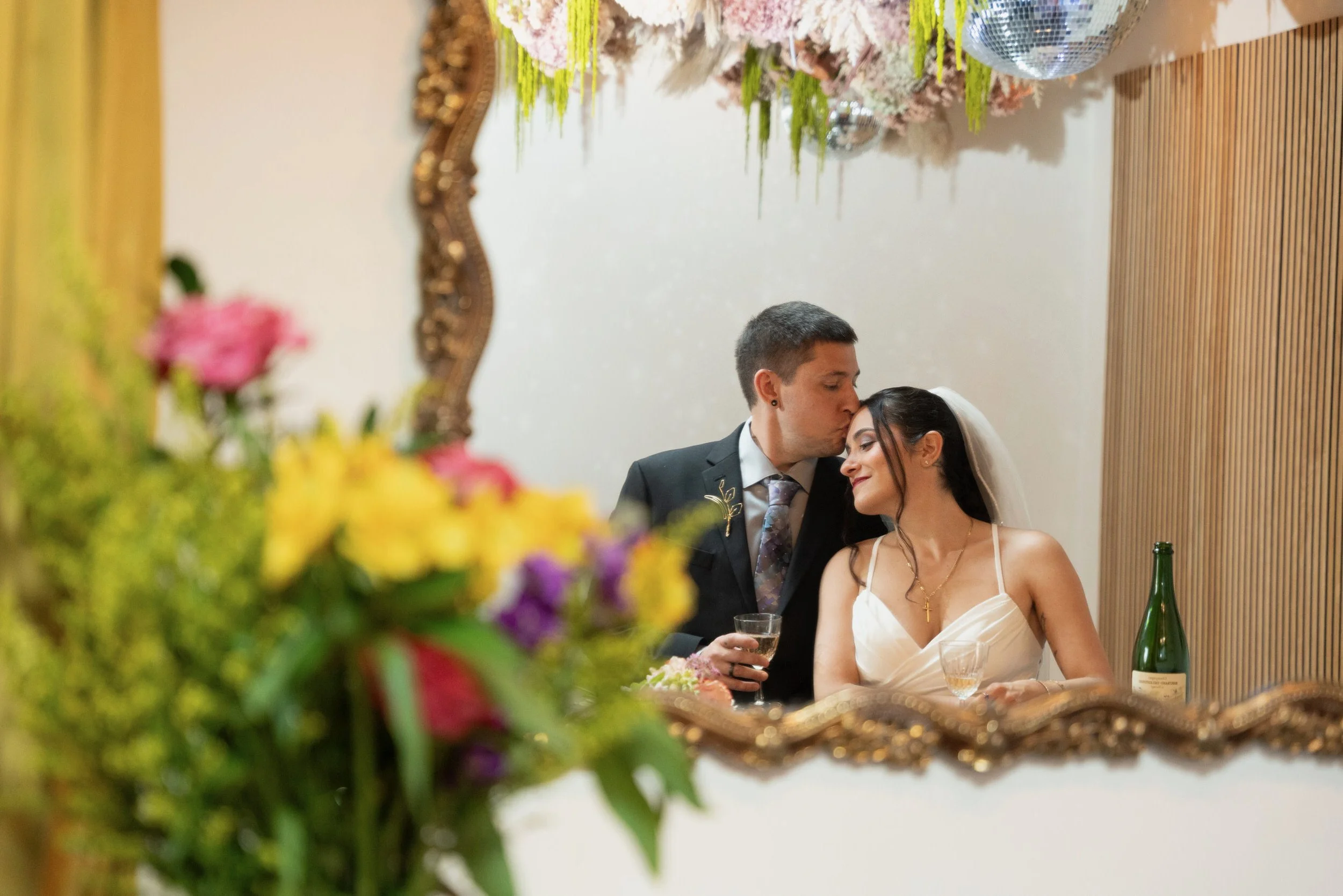 The reflection in the mirror of a newlywed kissing their partner's head