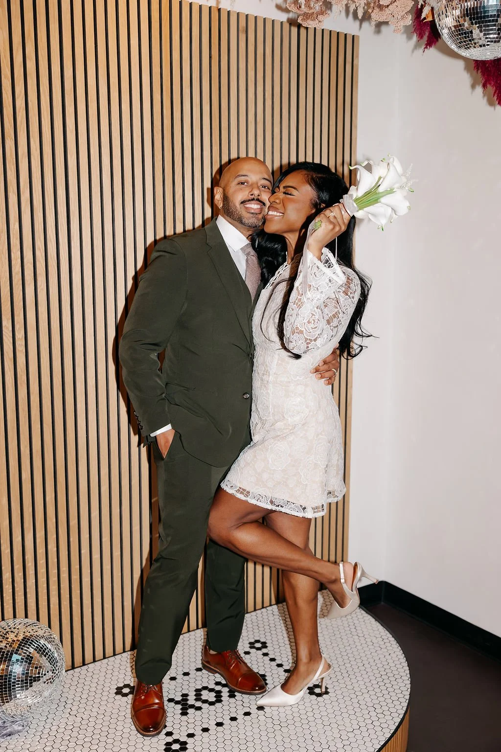 Couple standing on a tiled platform, looking as if they were just married. Both are smiling, one person is holding a bouquet of white flowers. 