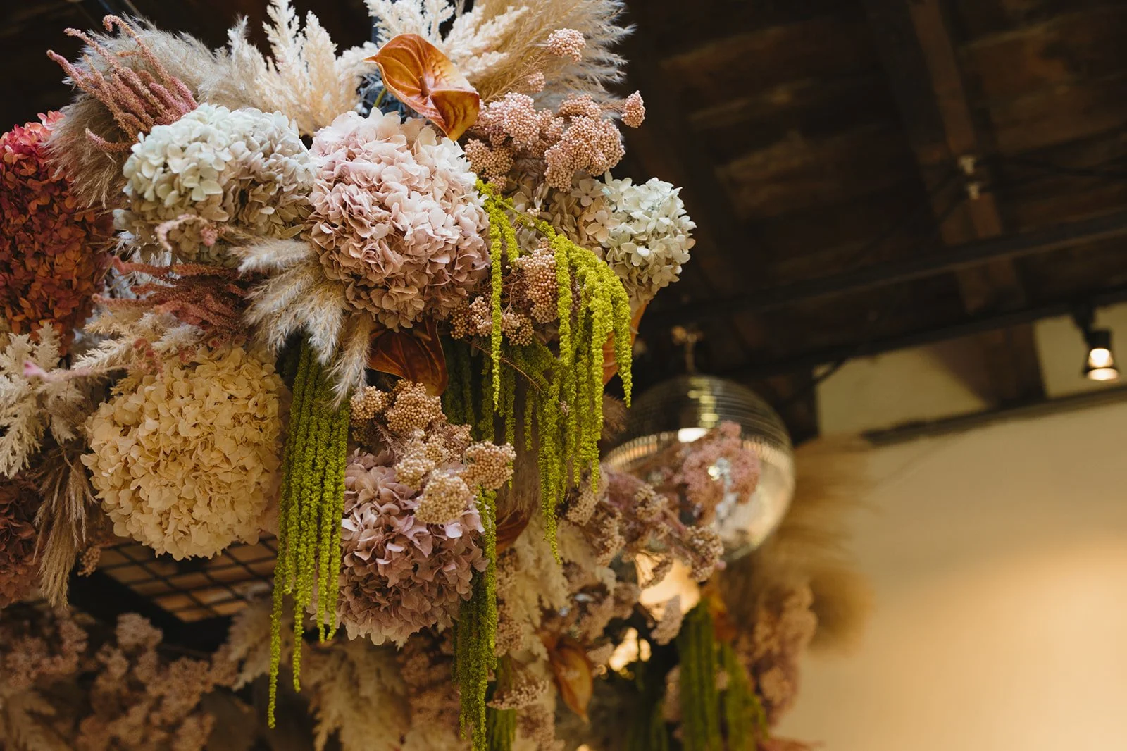 A floral arrangement with pink, cream, and brown hydrangeas, green hanging amaranthus, and other dried and fresh flowers hanging from a ceiling with wooden beams. A disco ball is in the background.