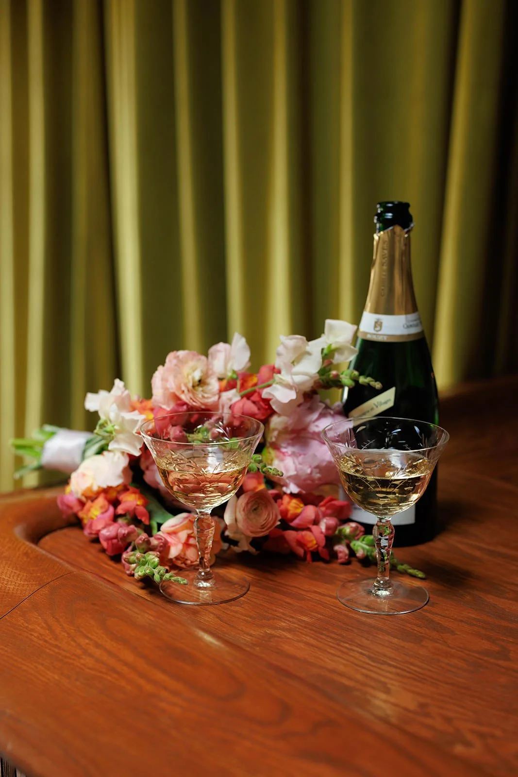 A bottle of champagne, two glasses of champagne, pink and white flowers, and a green curtain background on a wooden table.