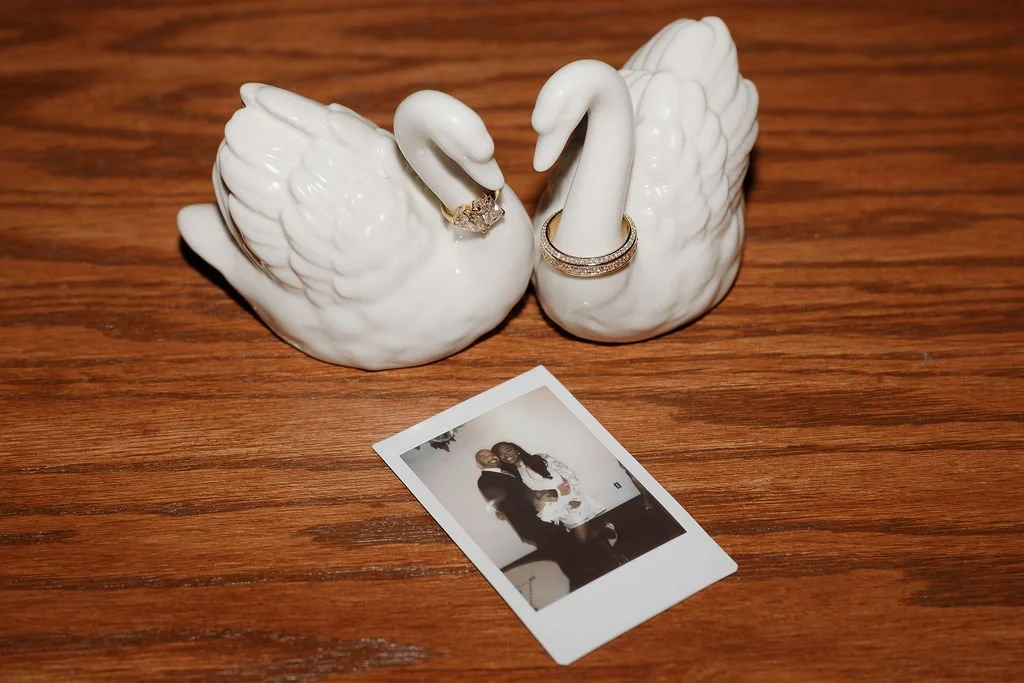 A ceramic swan-shaped jewelry holder with two rings displayed on its neck and a gold ring with diamonds on its head, placed on a wooden surface with a small photograph of two women in the foreground.