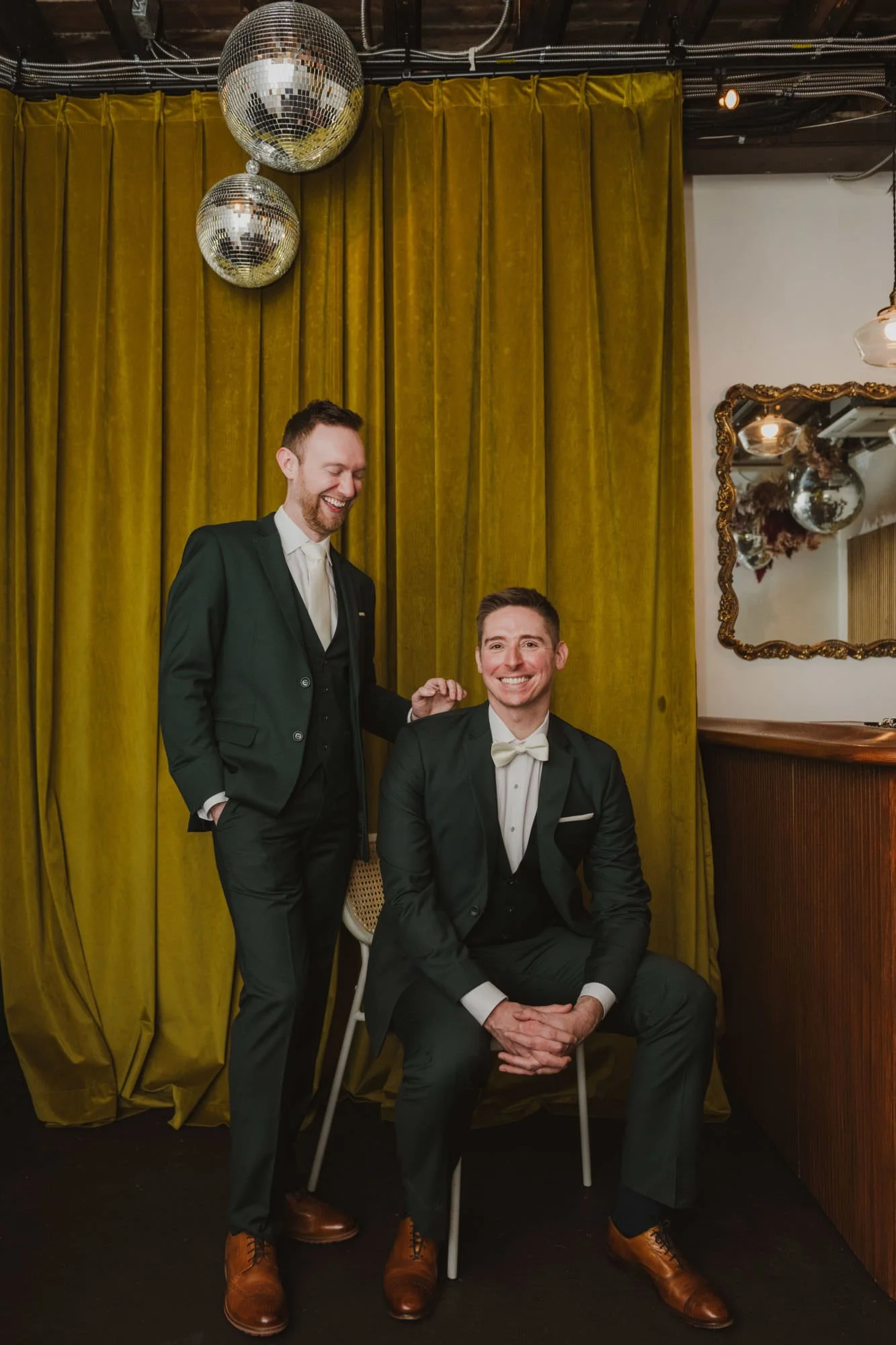 A newlywed sitting in a chair smiling with their partner standing next to them looking down at them while laughing