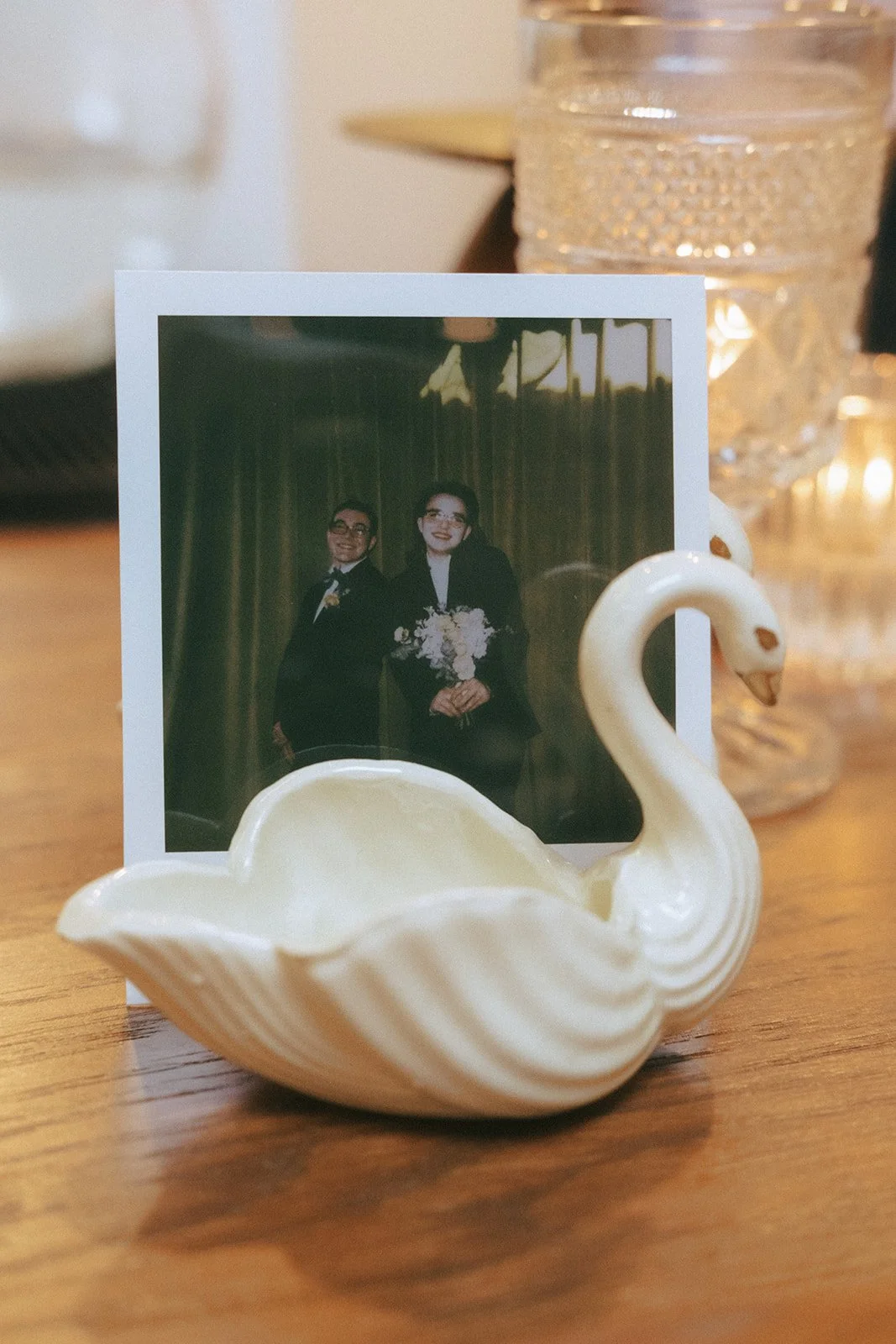 A small decorative swan-shaped dish holding a photograph of a happy couple in formal attire, with the man in a tuxedo and the woman in a black dress holding a bouquet of flowers, set on a wooden surface with candles and a glass container in the background.