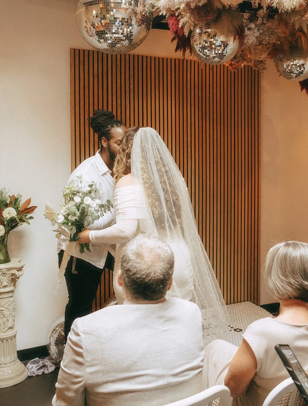 A wedding ceremony between two people, one with curly hair and the other with dreadlocks, facing each other, holding a bouquet, with guests watching. Discosphere lighting and floral decorations are visible.