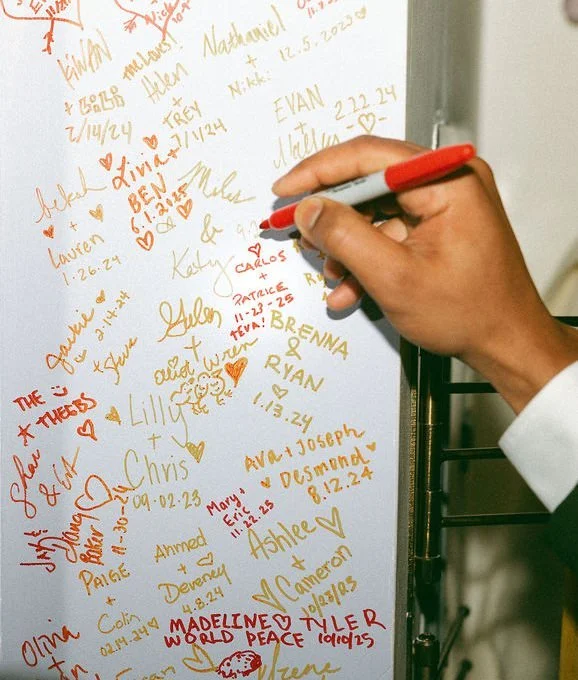 A hand signing a wall filled with couple's names and their wedding dates
