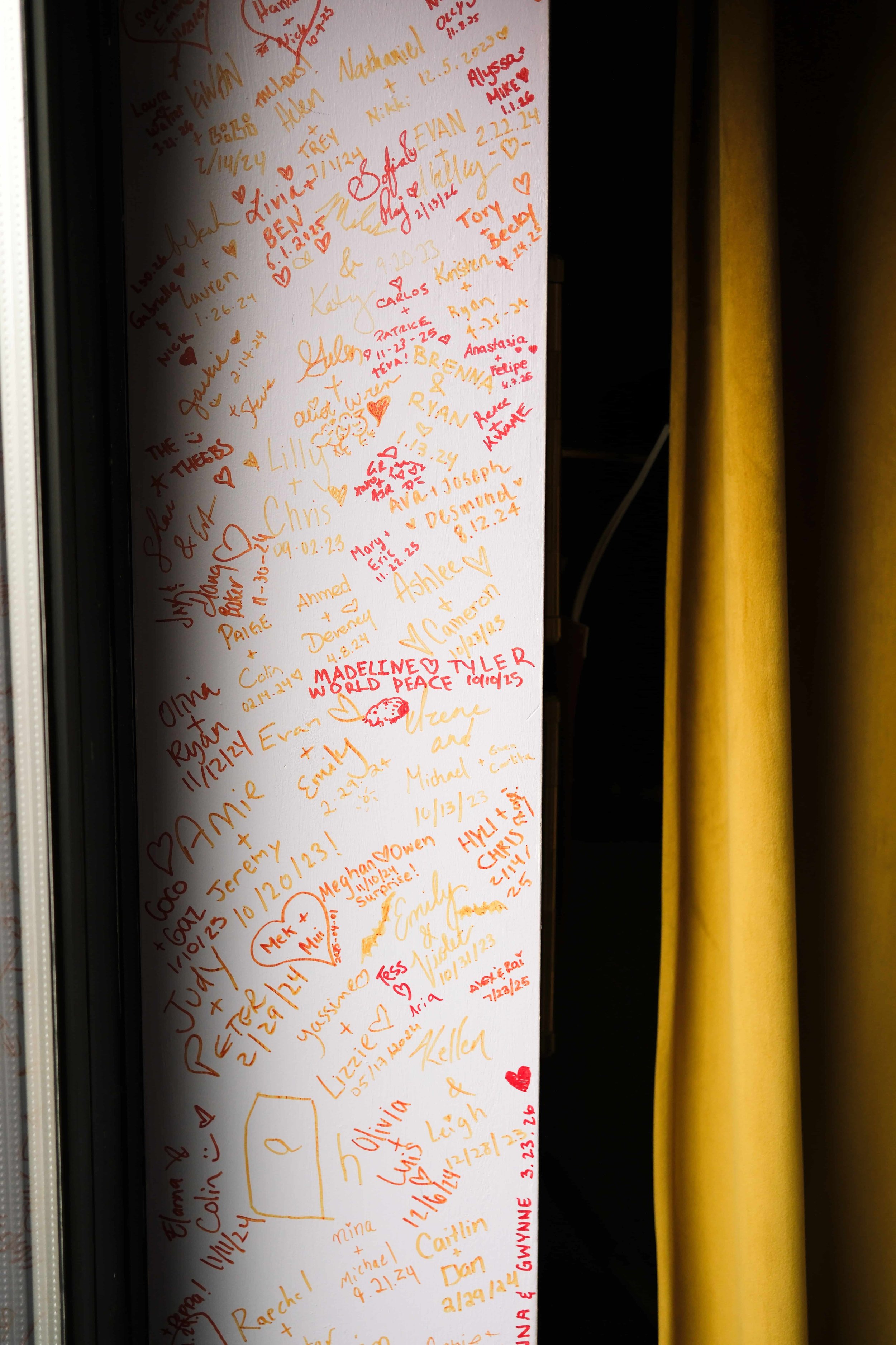 A white panel covered with colorful handwritten messages, signatures, and doodles in red, orange, yellow, and black ink, alongside small hearts and drawings. The messages appear to be celebratory, including names, dates, and well wishes.