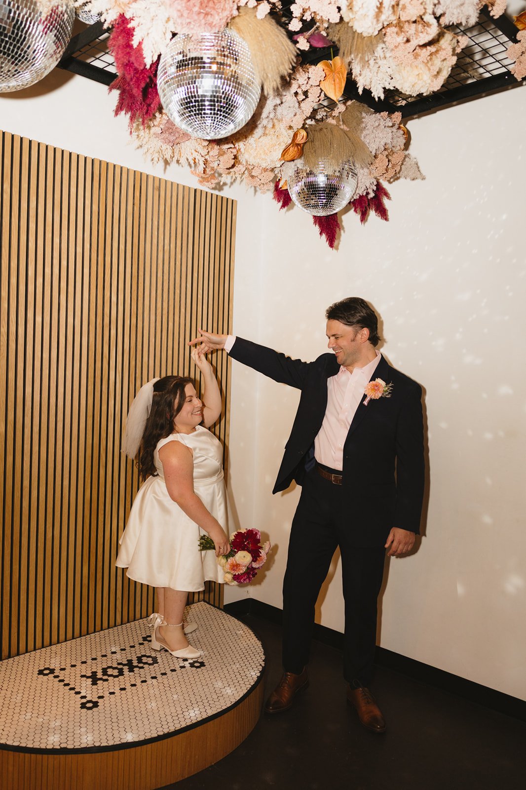 A man and a girl dancing together at a wedding, with the girl standing on a small platform holding a bouquet, while the man twirls her. The girl wears a white dress and veil, and the man is in a dark suit. The ceiling is decorated with pink and white