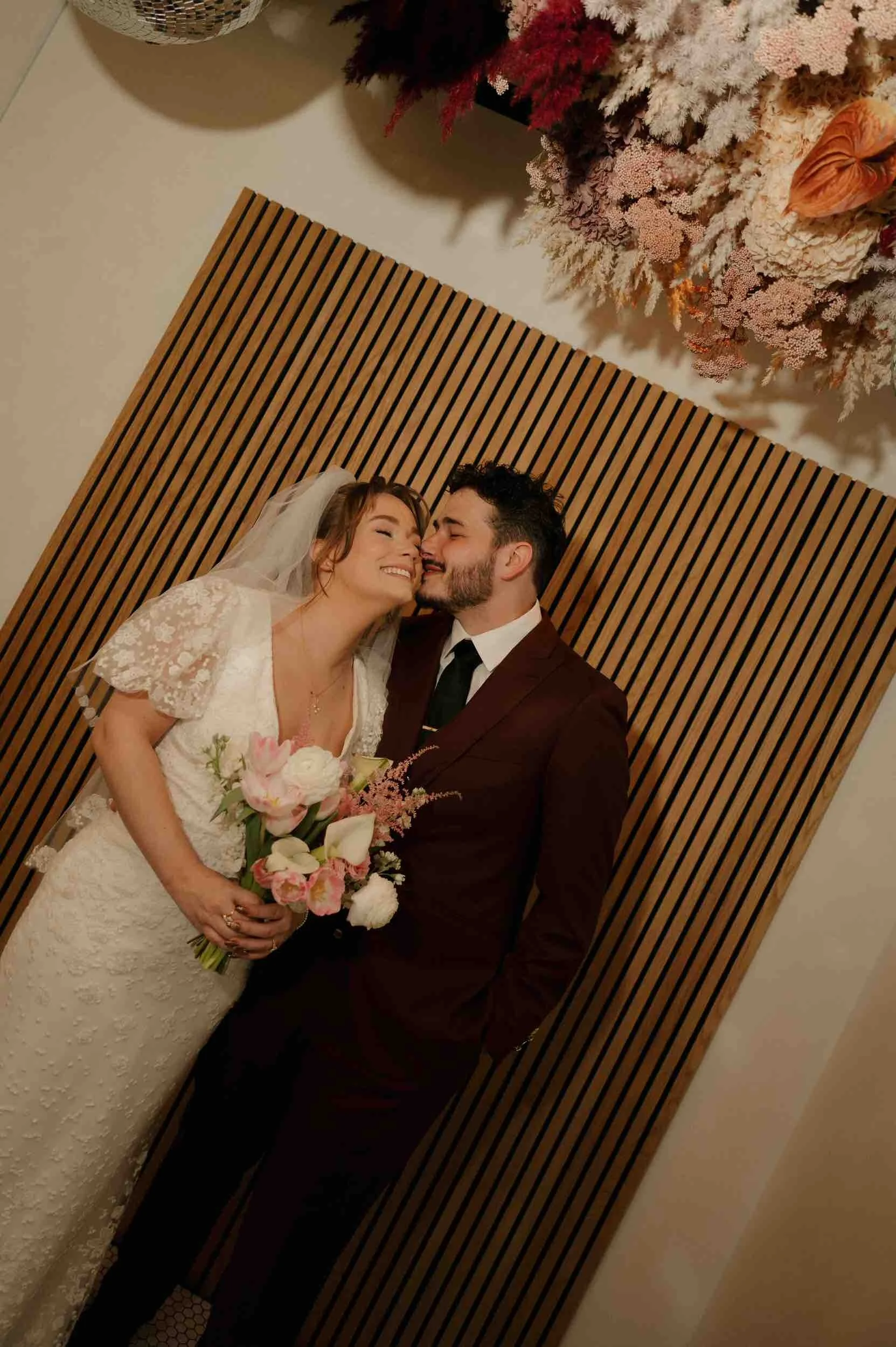 A newlywed leaning in to kiss their partner's cheek as they both smile