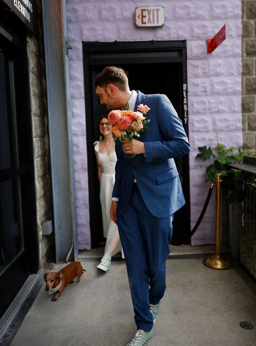 A man in a blue suit holding a bouquet of pink and orange flowers stands in front of a woman in a white dress, who is smiling as she peeks out from behind the doorway. A small brown dog stands near the door, and an exit sign is visible above.