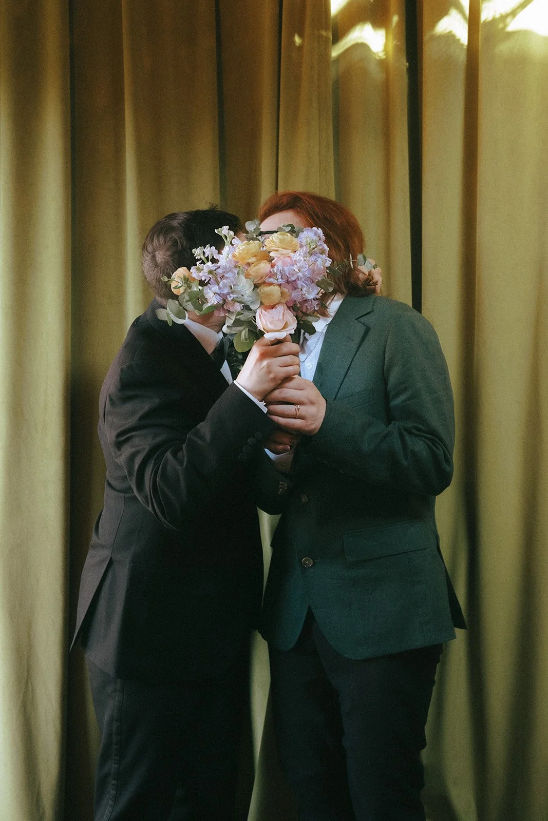 Two people in suits holding a bouquet of flowers, obscuring their faces, standing in front of a curtain.