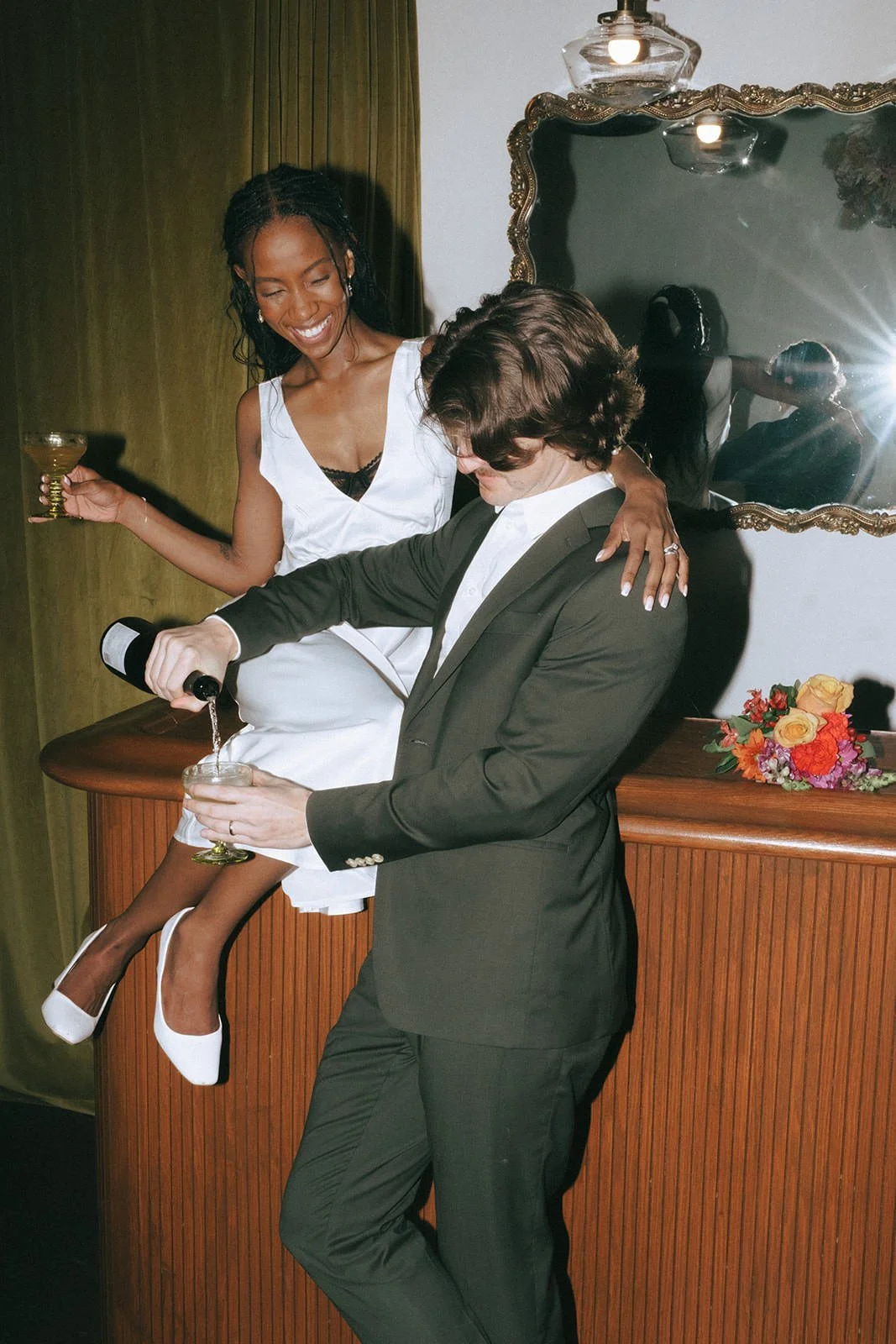 A person sitting on a cabinet with their arm around their partner's shoulder while their partner pours a drink