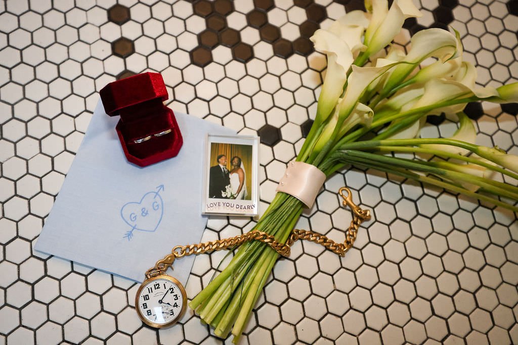 A bouquet of flowers, a polaroid, rings, jewelry, and a wedding invitation laid out on the ground