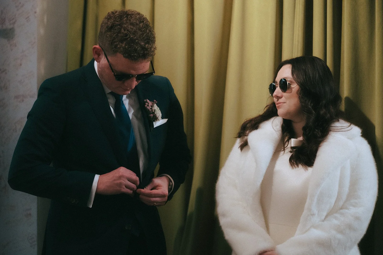 A man in a dark suit and sunglasses adjusting his jacket, and a woman in a white fur coat and sunglasses standing next to him, indoors with gold curtains.