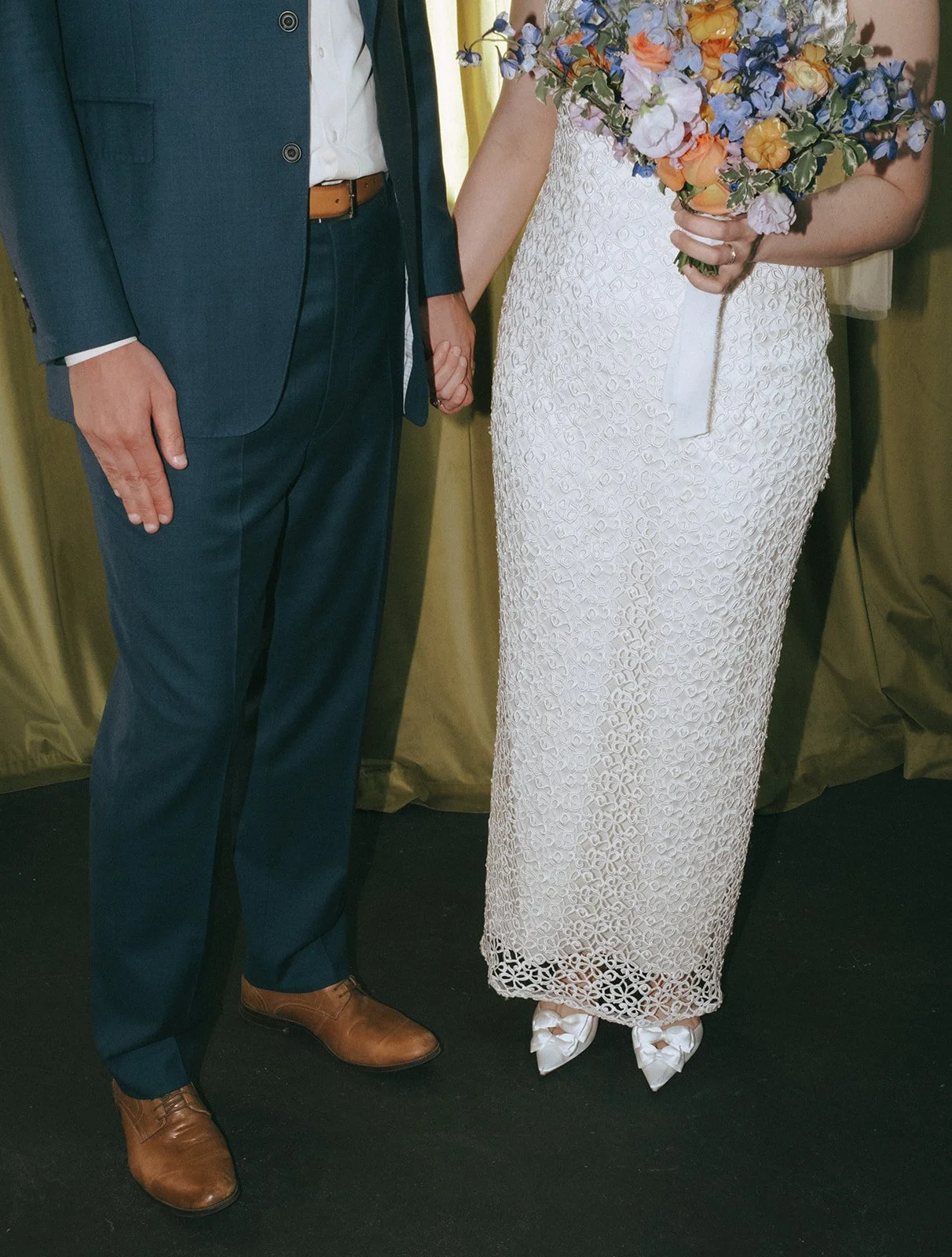 A couple holding hands, with the woman holding a bouquet of colorful flowers. The woman is wearing a white lace dress with satin shoes, and the man is dressed in a dark suit with brown shoes, standing on a black floor with a gold-colored curtain in the background.