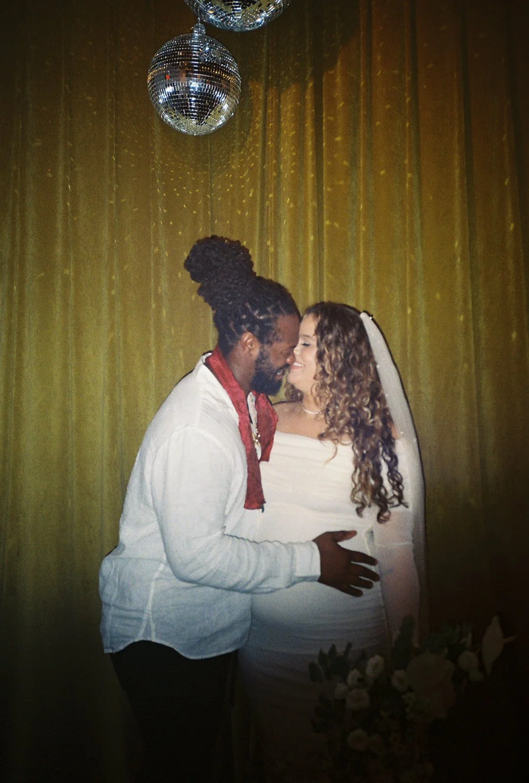 A couple smiling at each other, with disco balls and a fancy curtain in the background, dressed elegantly as if for a special occasion, possibly a wedding or celebration.