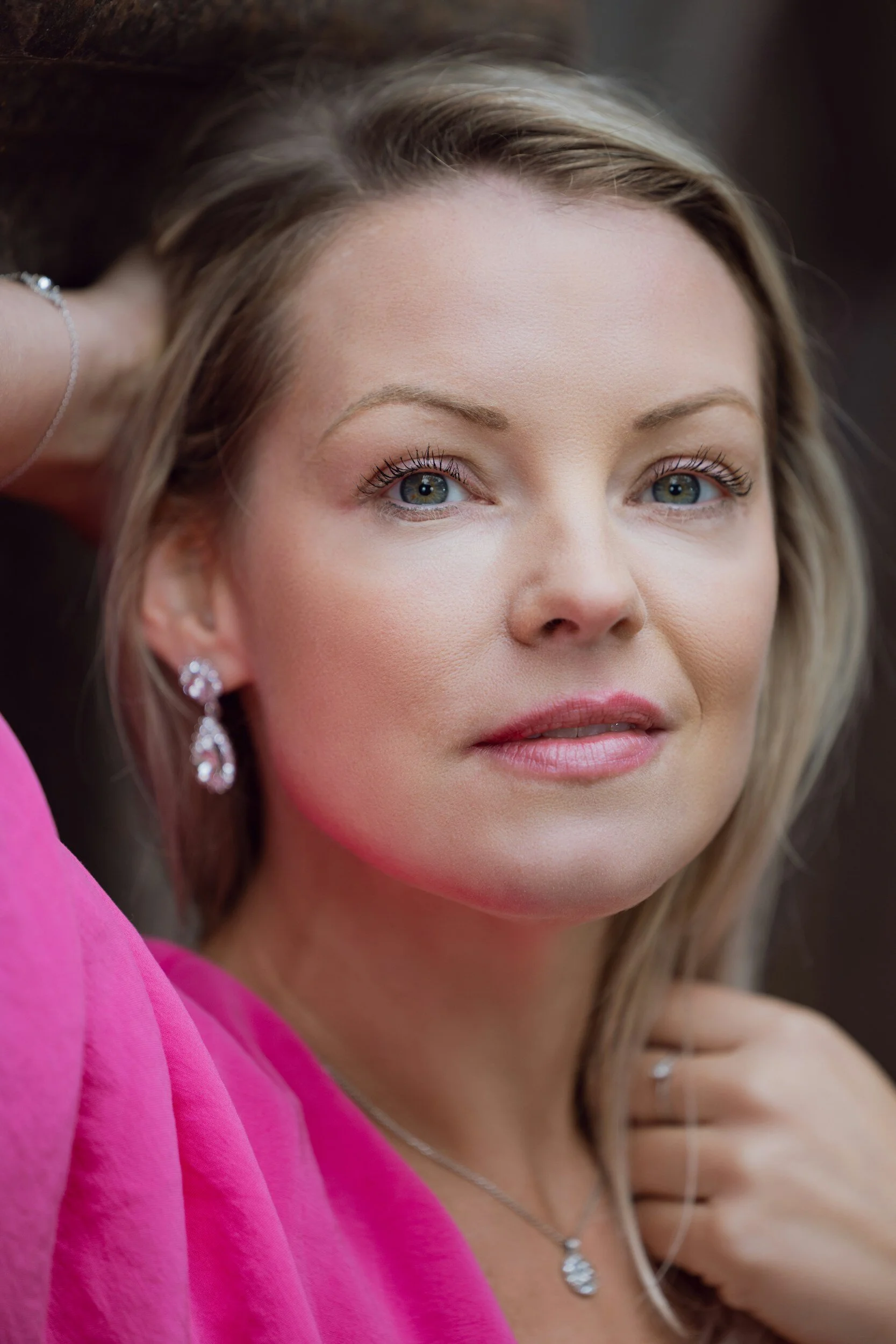Close-up portrait of a woman with blonde hair, blue eyes, and light makeup, wearing diamond earrings, a silver necklace, and a pink dress, looking into the camera with a soft expression.