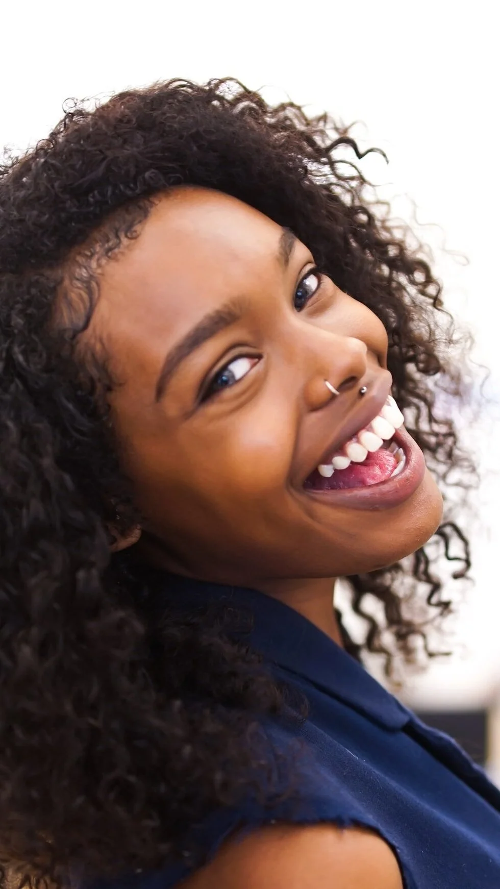 A young woman with curly hair is smiling and laughing, showing her teeth. She has multiple nose piercings and is wearing a dark blue top.