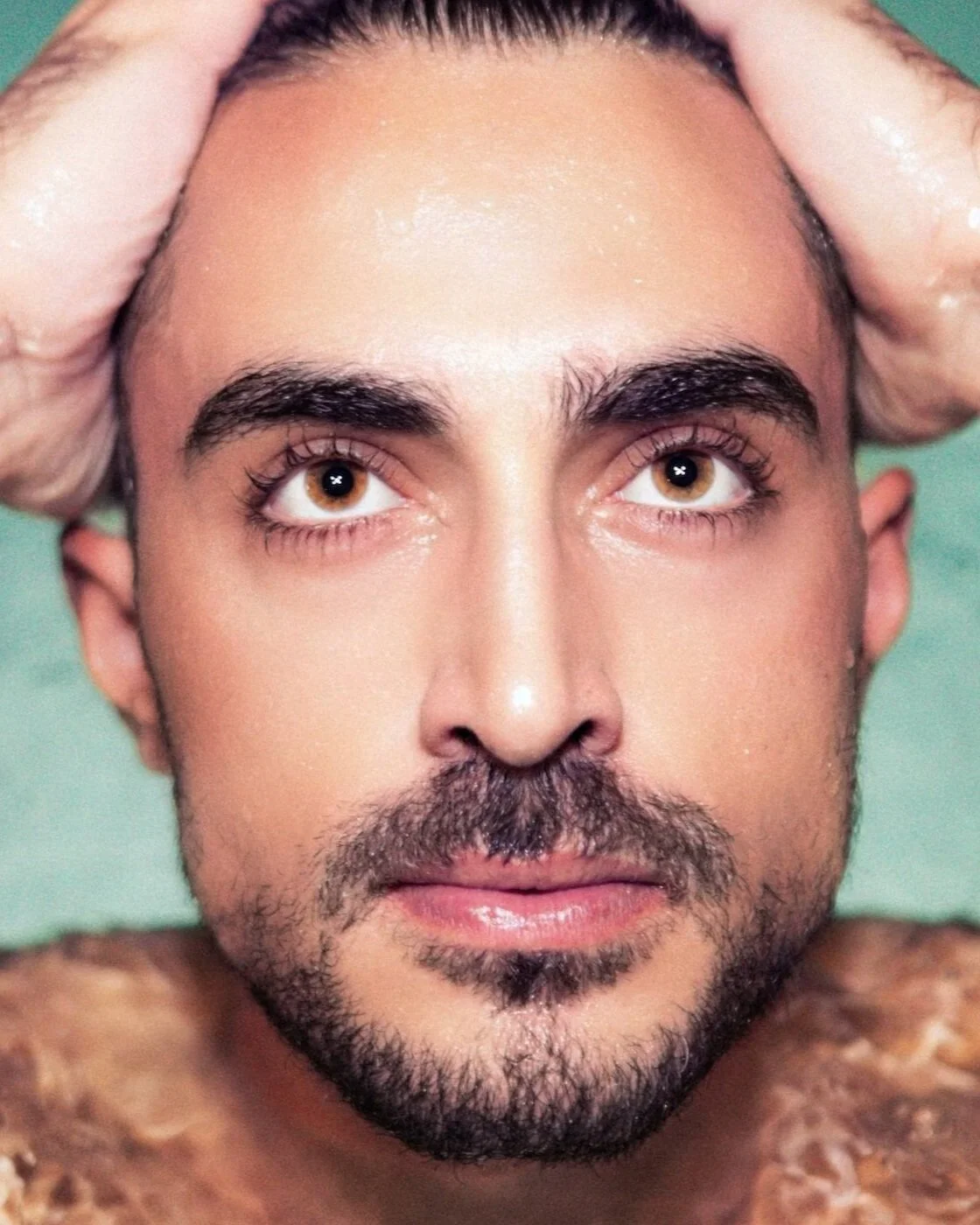 Close-up of a man's face with wet skin, brown eyes, dark eyebrows, facial hair, and hands on his head; background is light green.