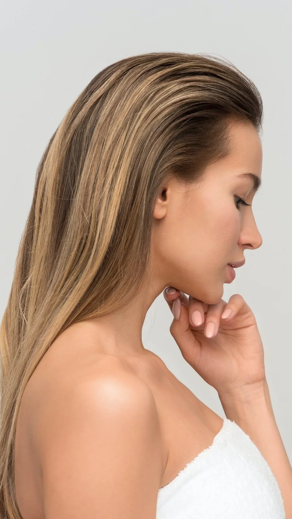 Side profile of a woman with long, straight, light brown hair, wearing a white towel, touching her chin with her hand, against a plain light gray background.