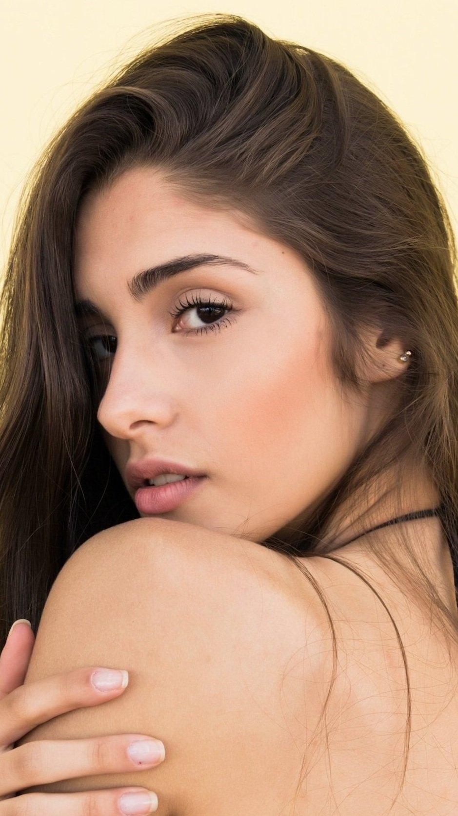 Close-up of a young woman with long brown hair, brown eyes, and light makeup, resting her hand on her shoulder.