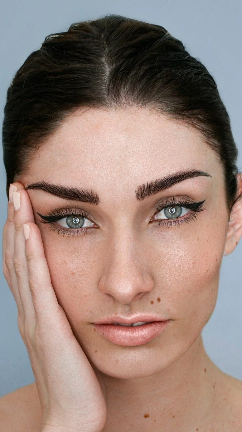 Close-up portrait of a young woman with blue eyes, dark eyebrows, and dark hair, touching her face with one hand, against a light blue background.