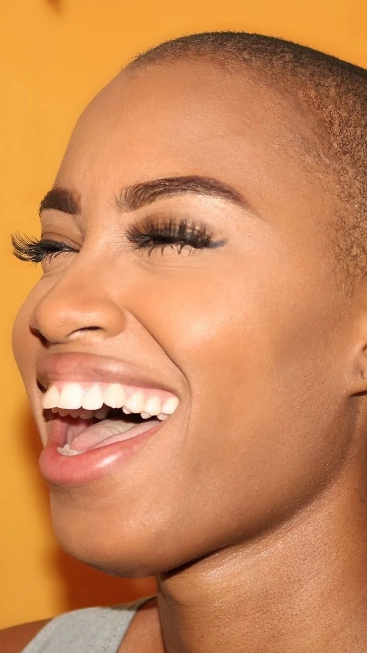 Close-up of a woman laughing with eyes closed, showing white teeth and wearing makeup, in front of a yellow background.