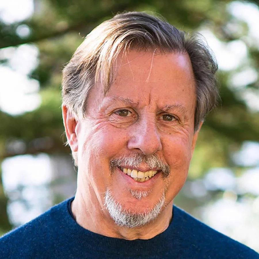 A smiling older man with gray hair, a mustache, and a goatee, wearing a dark blue shirt, outdoors with blurred trees in the background.