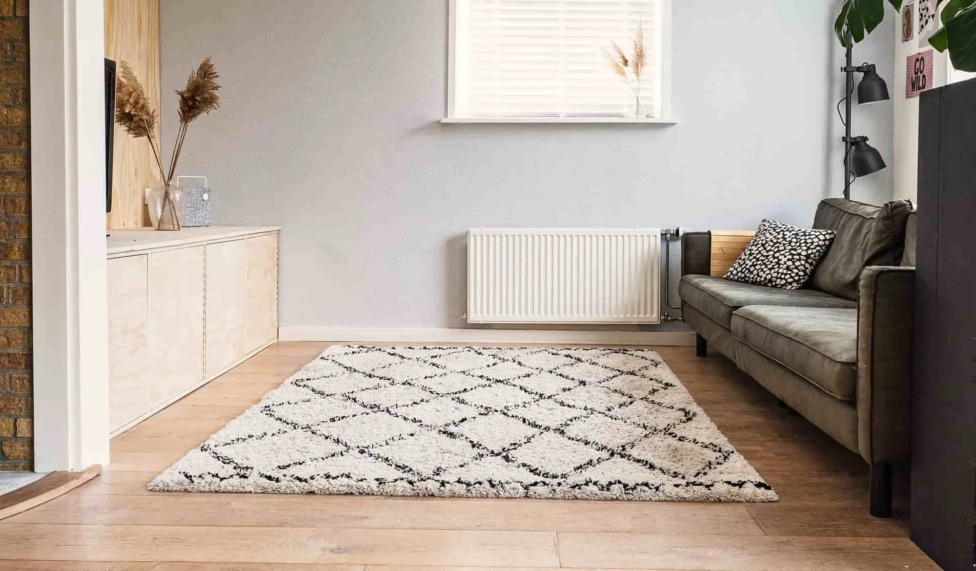 Living room with a white rug featuring black diamond pattern, gray sofa with patterned pillow, black wall-mounted lamps, and a wooden sideboard with decorative items.