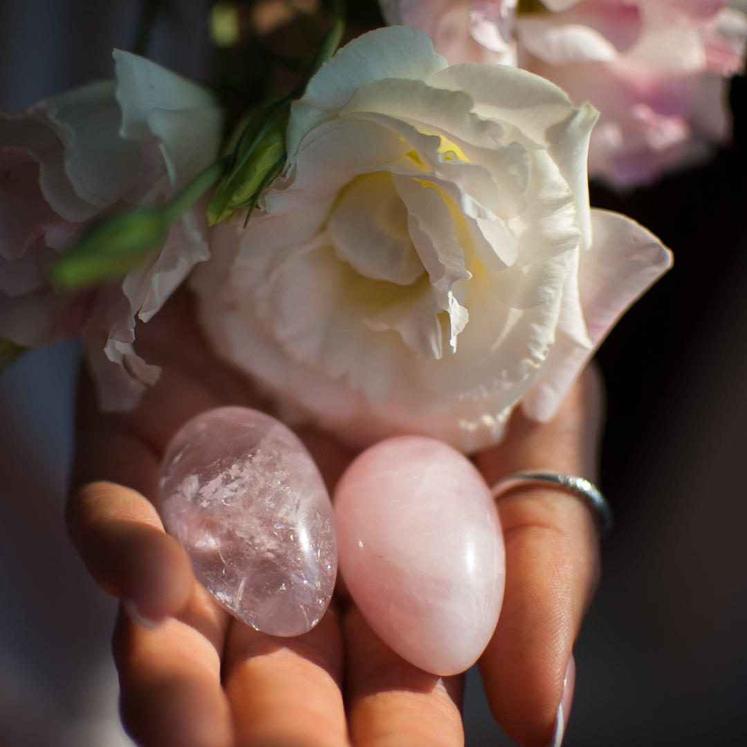 Close-up of a hand holding a white flower and two polished stones, one clear and one pink.
