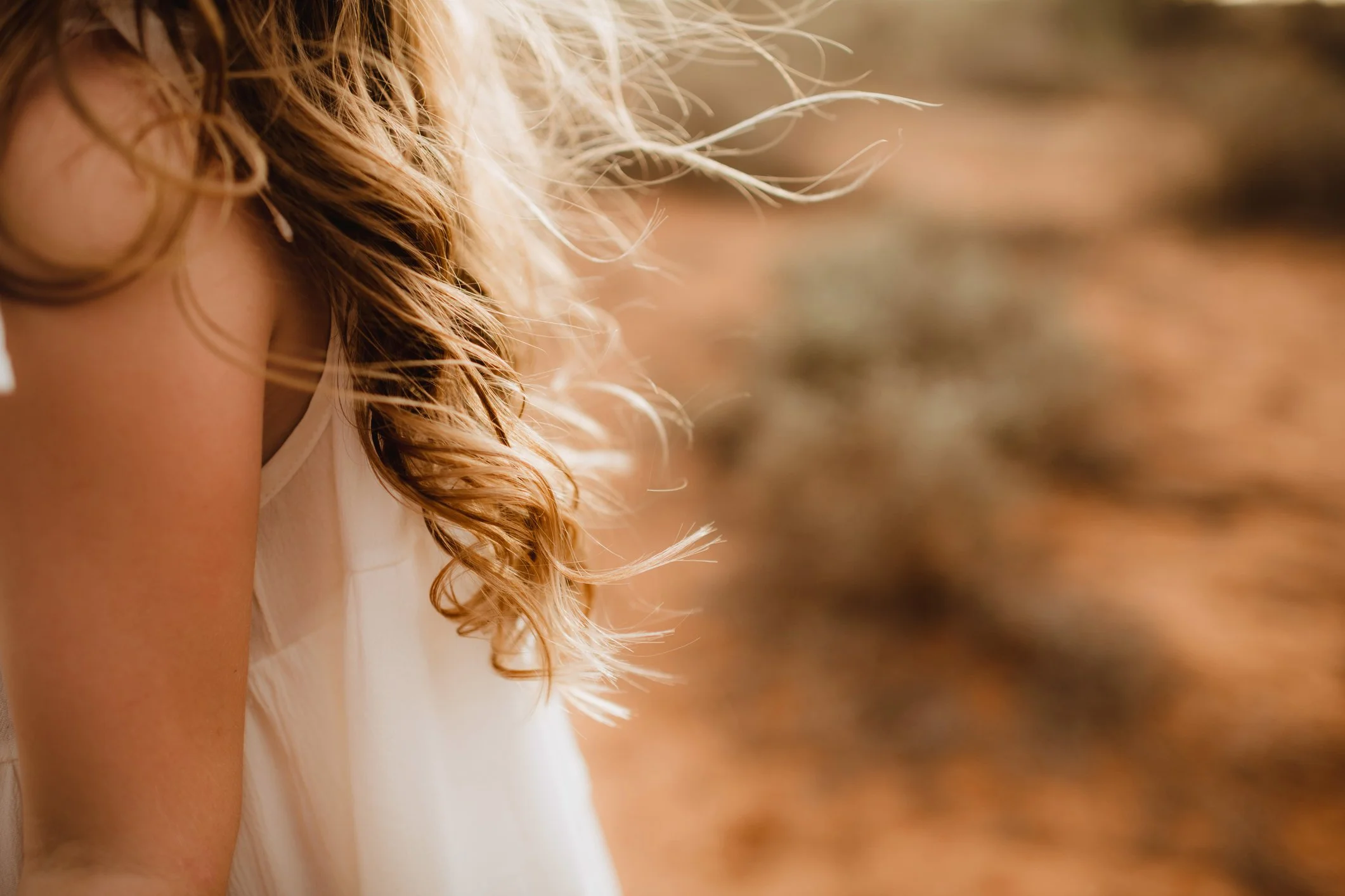 Close-up of a woman's shoulder and curly blonde hair, wearing a white sleeveless top, outdoors with a blurred natural background.