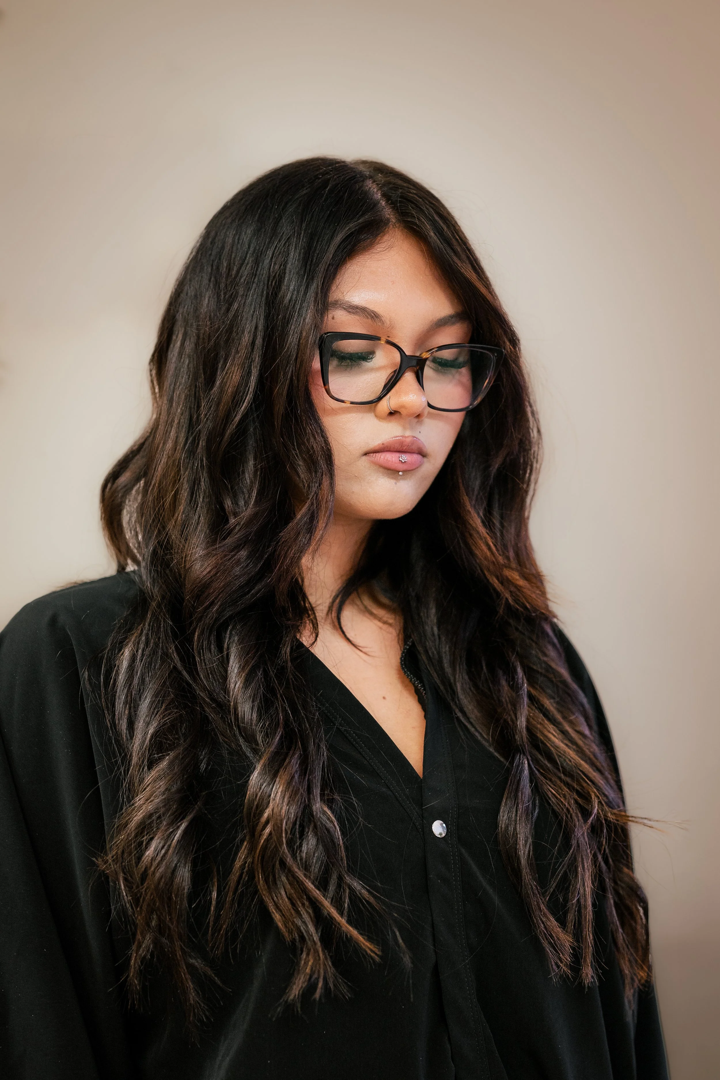 A young woman with long, wavy dark hair with purple tips, wearing large black-rimmed glasses, and a black button-up shirt, is looking downward with closed eyes against a plain light background.