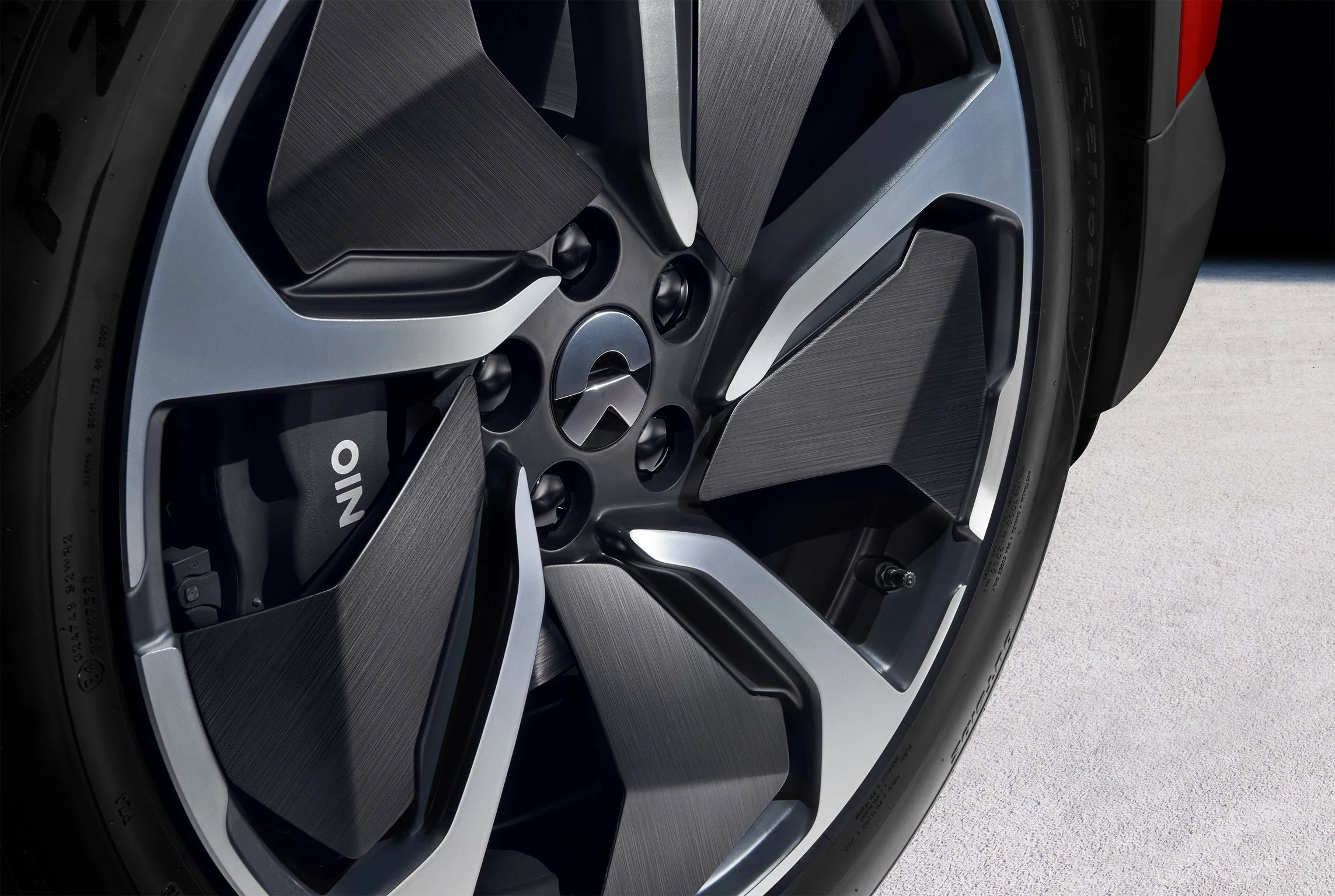 Große Automobilfelge mit einem modernen, angularen Design, das hauptsächlich in Grau und Schwarz gehalten ist. Im Zentrum befindet sich ein Logo, wahrscheinlich eines Automobilherstellers, und es ist ein Teil des Reifens sichtbar.