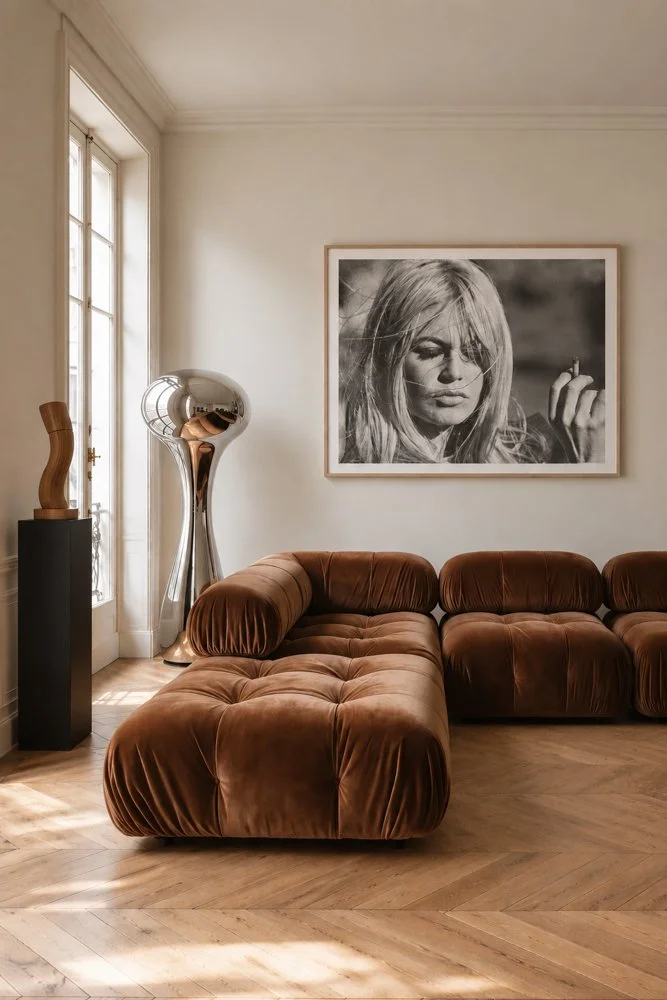 A living room with a curved brown velvet sofa, a tall metallic sculpture, a framed black-and-white photograph of a woman, a window letting in natural light, and hardwood flooring.
