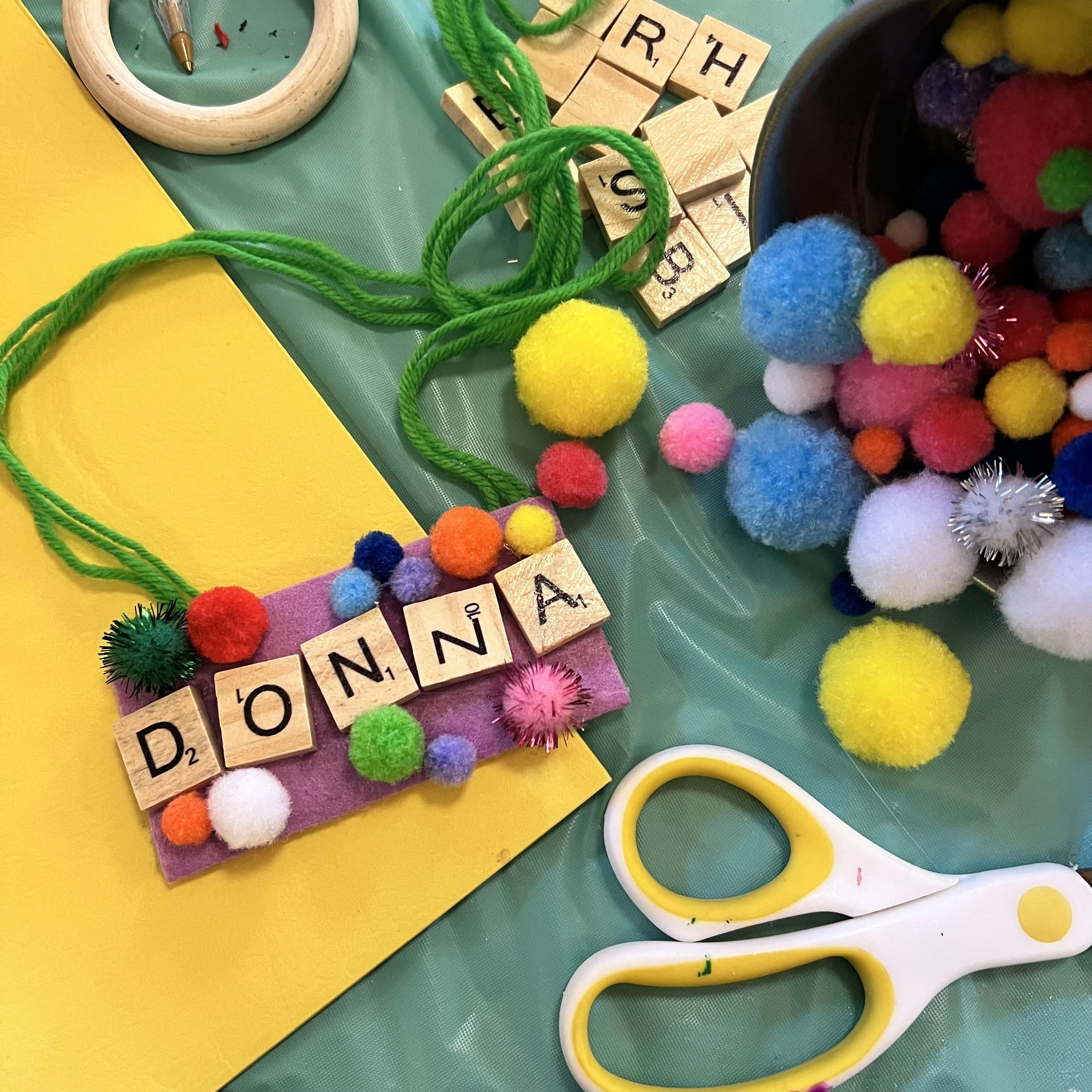 Colorful craft supplies on a green background and a playful name tag with pom poms and the name "Donna" spelled out in scrabble tiles. Around the name tag are scissors, assorted colors of pom poms, yellow paper, and scrabble tiles.