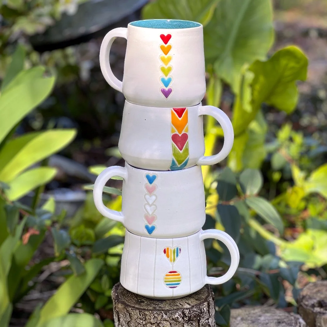 A stack of four mugs from Stuffed Studio's Pride collection. Matte white base with different glossy, colorful heart designs in Pride flag colors.