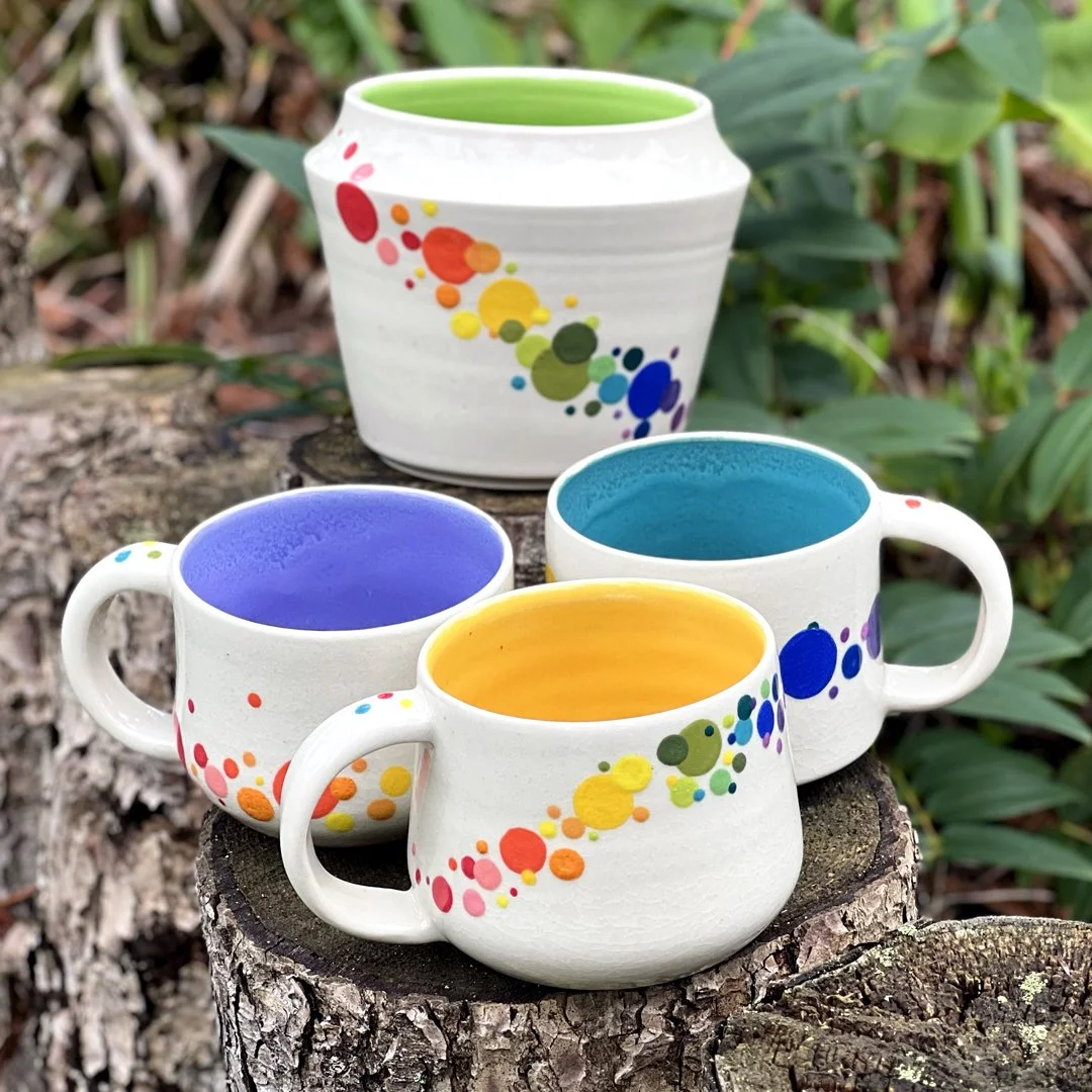 Three colorful mugs and one large vase from Stuffed Studio's Pom Pom collection. All feature a white, glossy clay with a rainbow ombré pattern of different sized dots. Each with a different bright color on the inside.