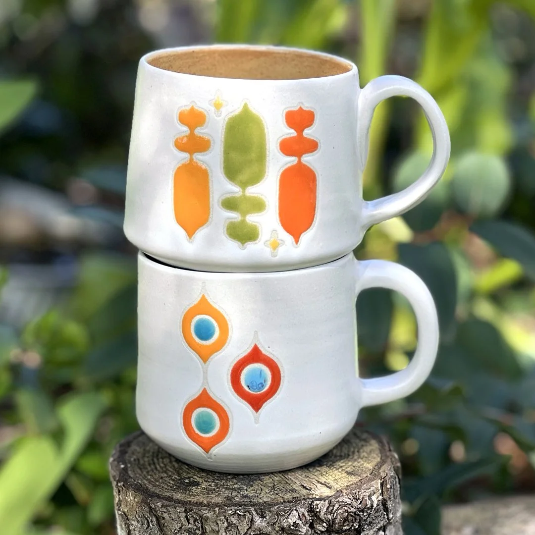 Two mugs stacked from Stuffed Studio's Retro Revival Collection. Both feature a matte white base with two different colorful mid century modern patterns in glossy oranges, greens, golds, and turquoise.