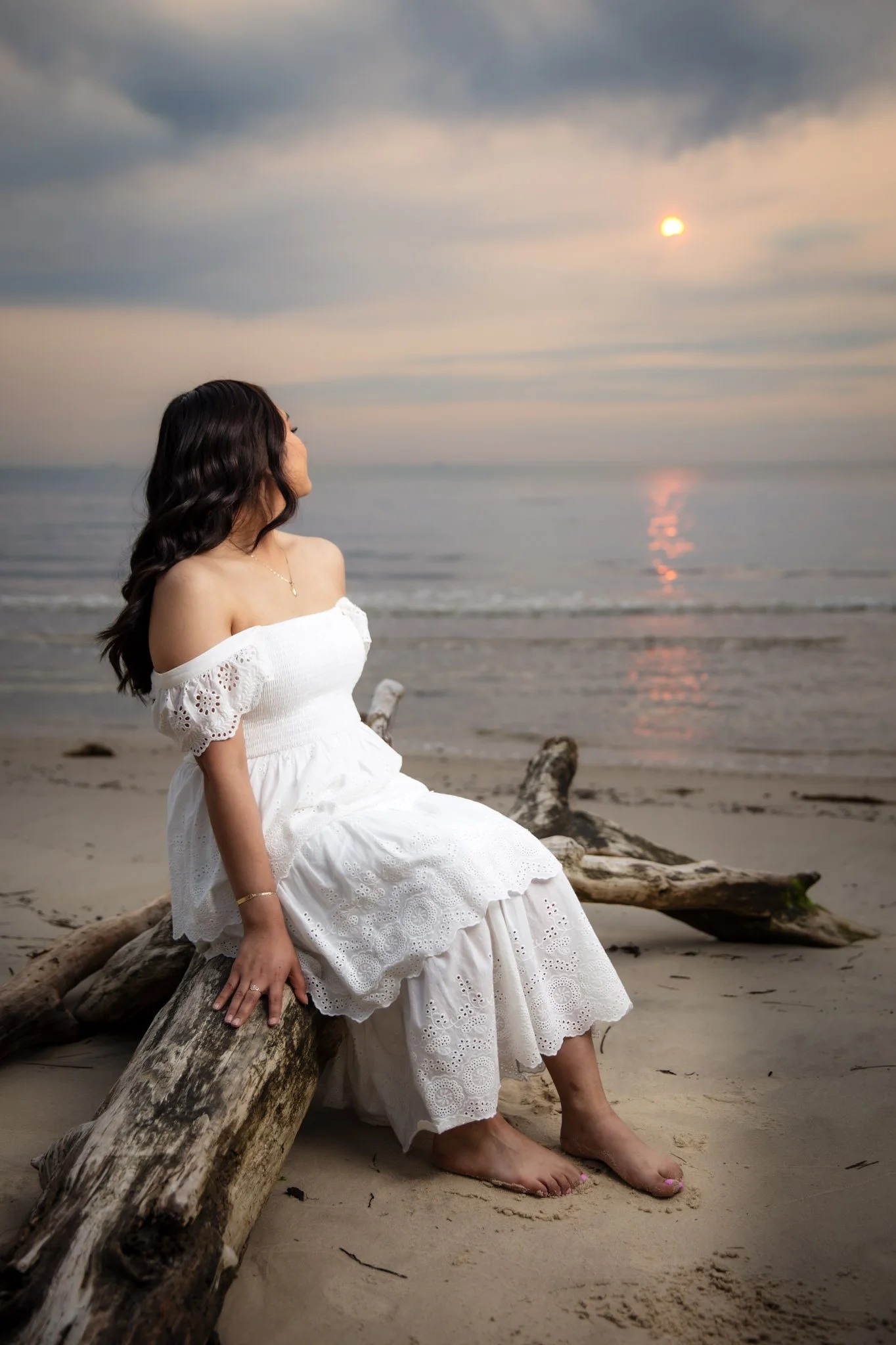 A woman in a white dress sitting on a piece of driftwood on a sandy beach at sunset.