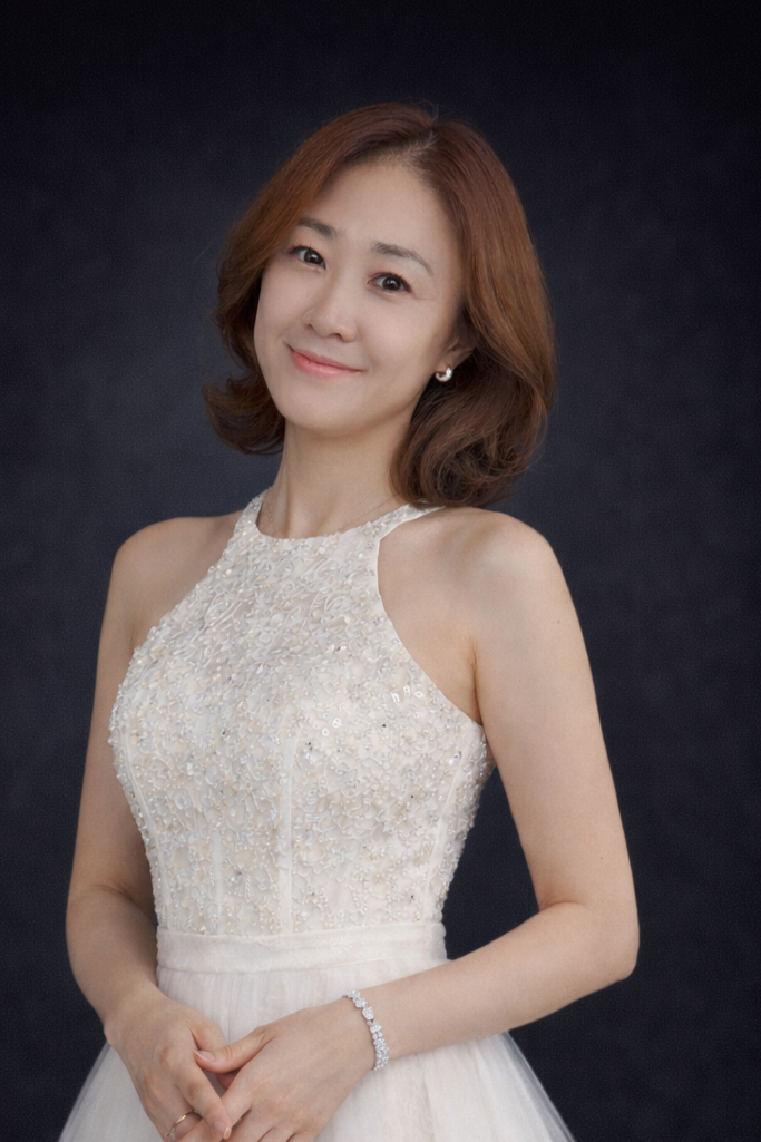 A woman with shoulder-length brown hair, wearing a white, lace-embellished sleeveless dress, smiling softly, posed against a dark background.