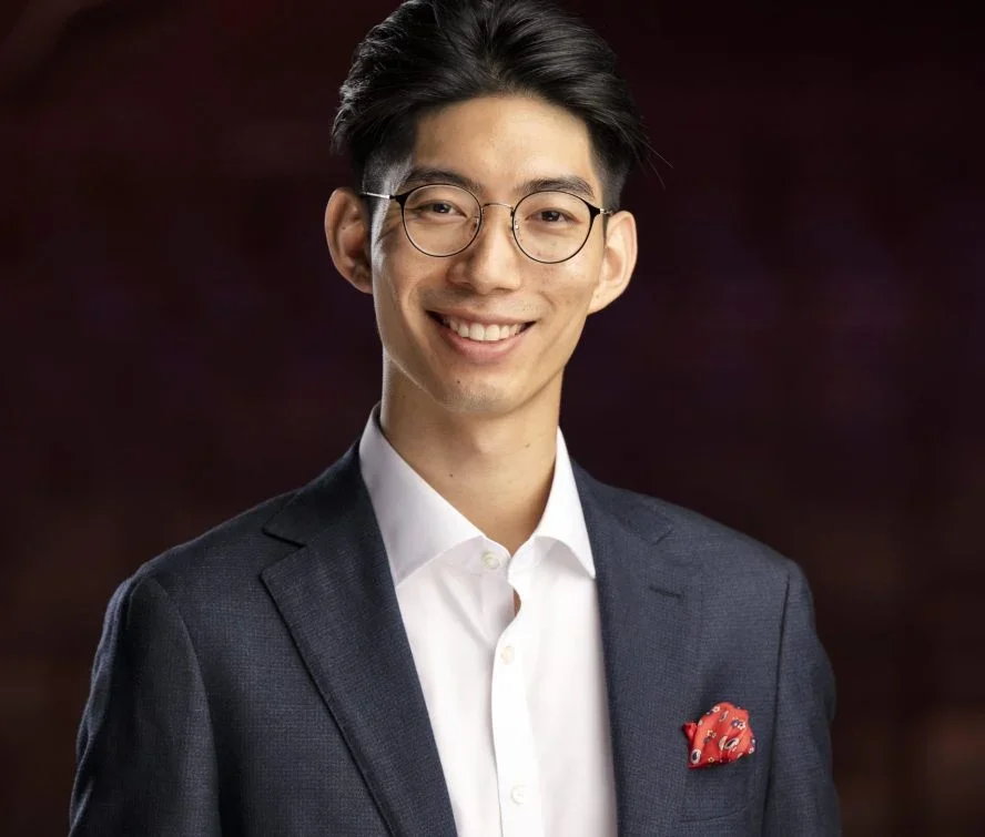 A young Asian man with glasses, wearing a dark suit, white shirt, and red pocket square, smiling confidently against a dark blurred background.