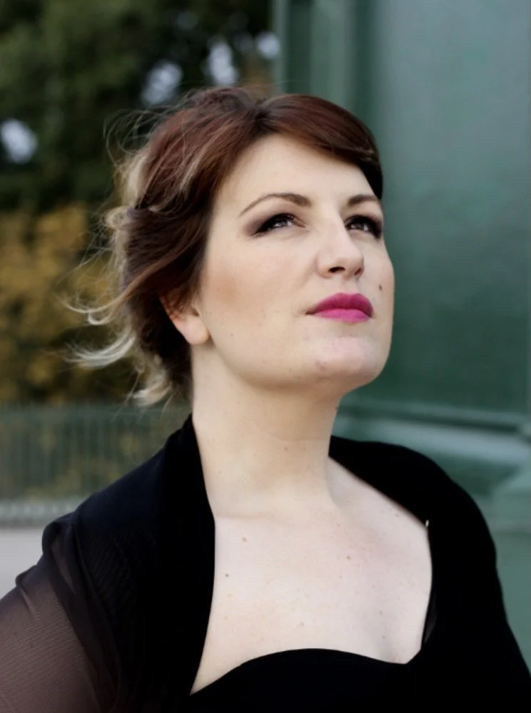 A woman with short reddish-brown hair, pink lipstick, and a serious expression, standing outdoors near a green structure.
