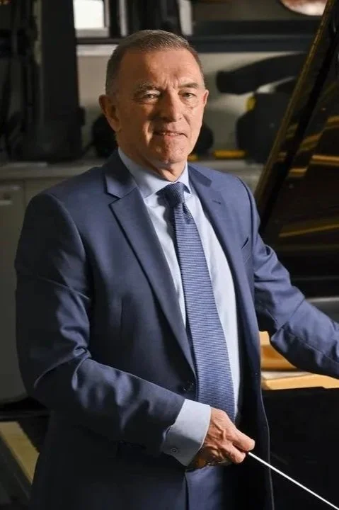 A man in a blue suit standing next to a grand piano, holding a conductor's baton, in a music rehearsal space.