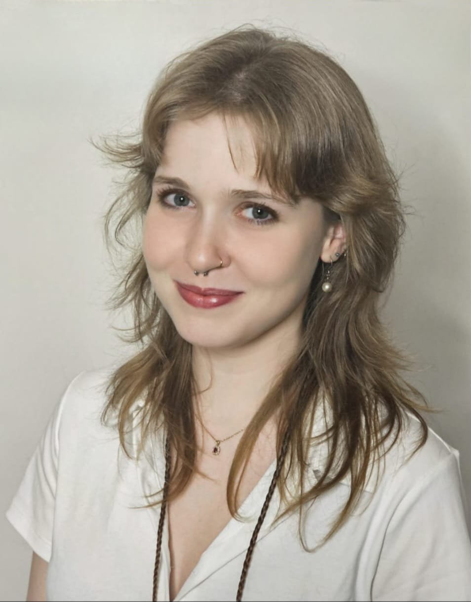 A woman with shoulder-length red hair, wearing a patterned shirt, smiling and looking at the camera against a plain background.