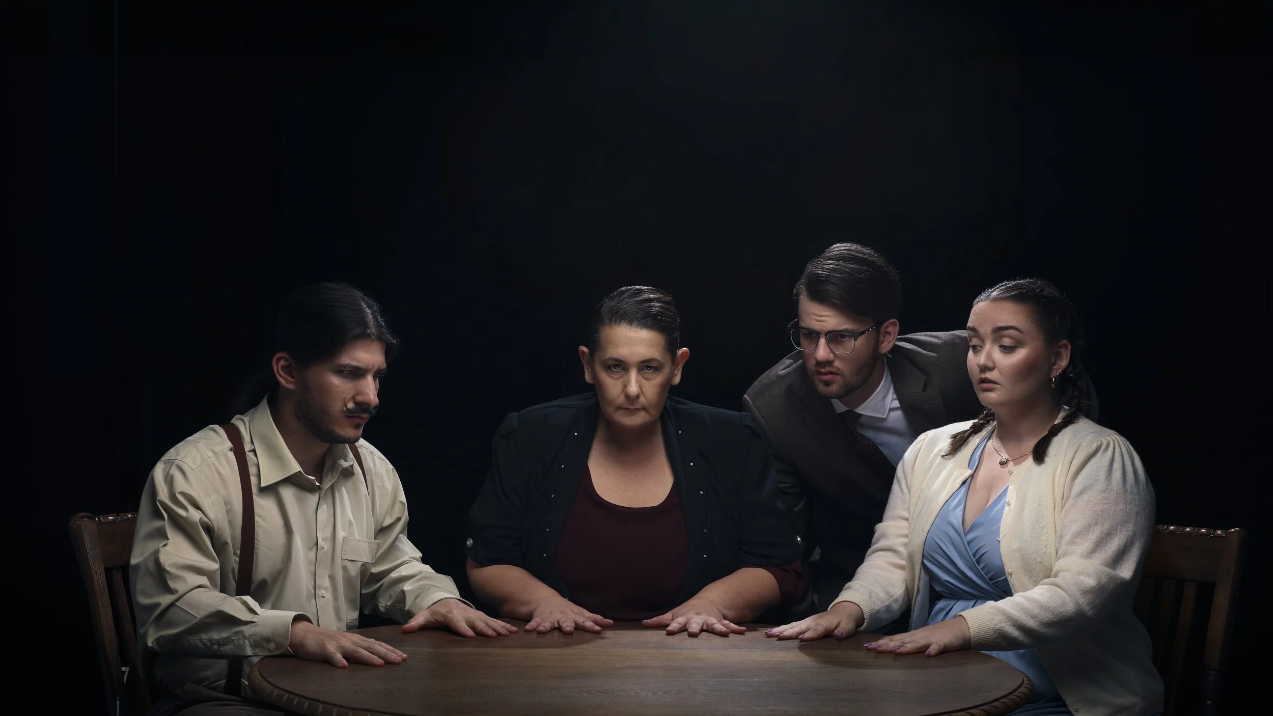 Four people sit around a dark wooden table with serious expressions, against a black background.
