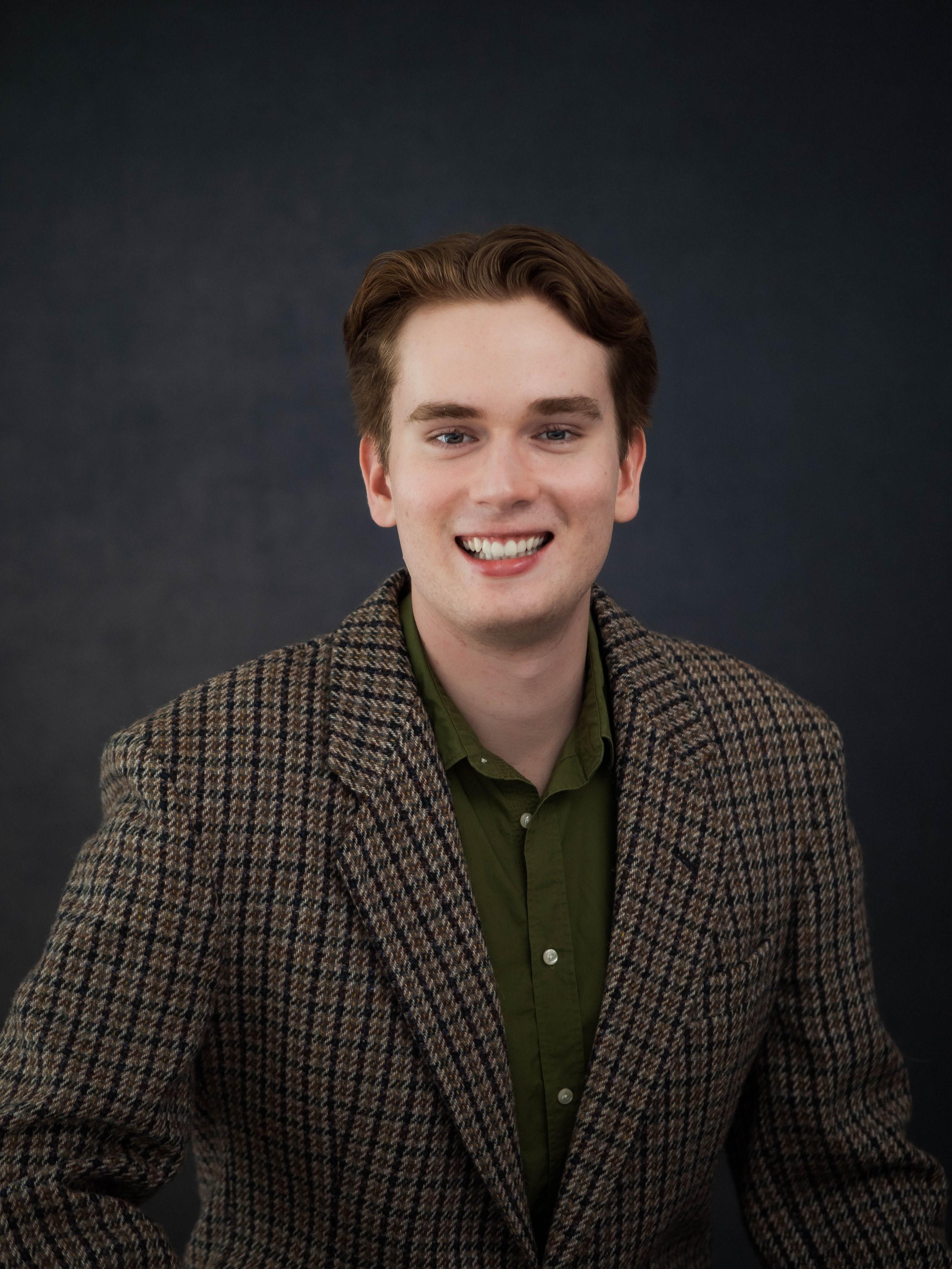 A young man with brown hair, wearing a green shirt and a brown checked blazer, smiling at the camera against a dark background.