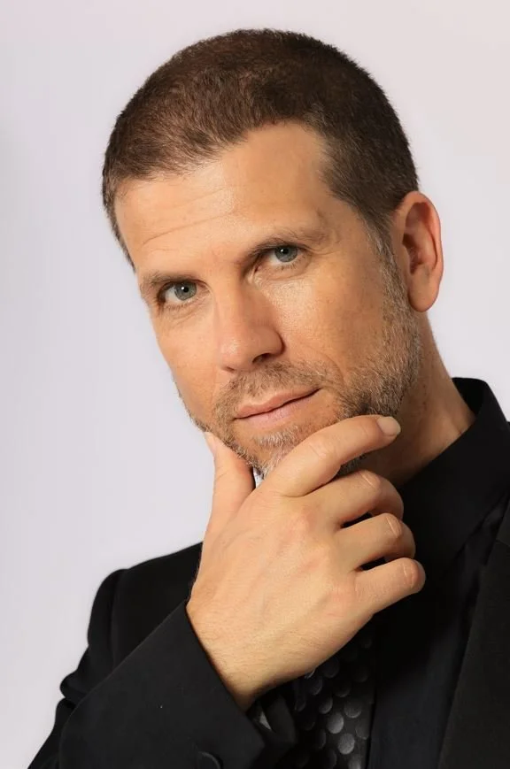 A man with short brown hair and blue eyes, dressed in a black suit, is touching his chin with his right hand and looking at the camera against a plain light background.